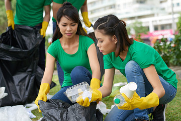 Two pretty female friends sitting on haunches while picking up plastic household waste in park