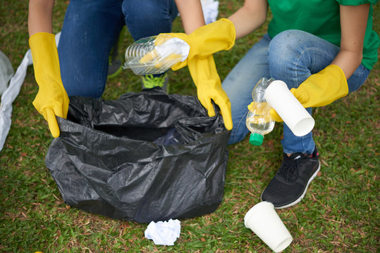Young woman holding black bin bag while her friend putting plastic household waste into it, close-up shot
