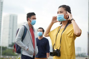 Waist-up portrait of Asian woman putting on protective mask while standing in city center and looking into distance