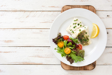 Fried cod fillet and salad in plate on white wooden background
