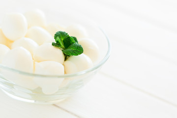 Small balls of mozzarella in glass bowl decorated fresh mint leaves on white wooden table. Selective focus