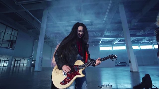 Young emotional expressive asian musician woman in black leather jacket playing the electric guitar in hangar.