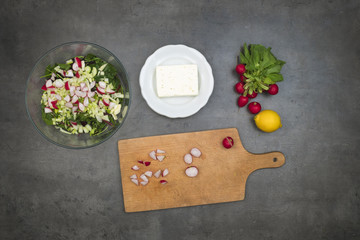 Preparing fresh vegetable salad with greek cheese. Top view on grey kitchen counter with ingredients and accessories. Food background.