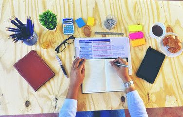 Top view of A young Asian woman's hands that writing on notebook with Smartphone wooden table background
