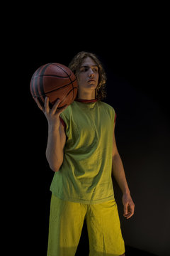 Young boy with long blond hair wearing a green jersey standing and holding up a basket ball