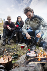 Man Cooking Food On Campfire With Friends In Background