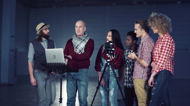 Bold man producer with laptop talking to crew of young mixed ethnic people filmmakers standing near DSLR camera on tripod in dark empty hall