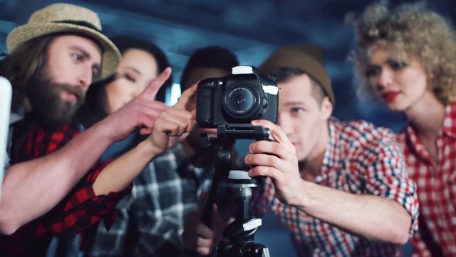 Group of young diverse people filmmakers gathered behind DSLR camera on tripod to check the angle of shooting in studio. Pointing fingers and smiling