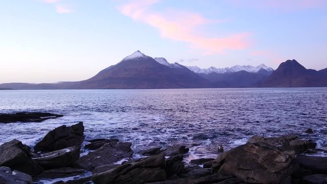 Evening in Elgol bay on west coast of the Isle of Skye in Scotland during an windy sunset . Snowy mountains on Soay island above sea of the Hebrides, waves crashing against to boulders and sharp rock 