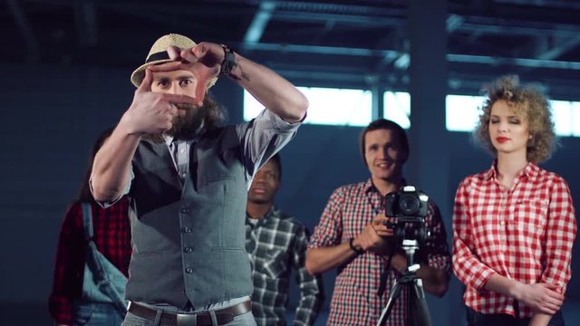 Young bearded man in hat stepping out and making frame gesture with fingers while group of young filmmakers shooting video with dslr camera