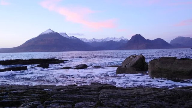 Evening in Elgol bay on west coast of the Isle of Skye in Scotland during an windy sunset . Snowy mountains on Soay island above sea of the Hebrides, waves crashing against to boulders and sharp rock 