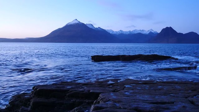 Evening in Elgol bay on west coast of the Isle of Skye in Scotland during an windy sunset . Snowy mountains on Soay island above sea of the Hebrides, waves crashing against to boulders and sharp rock 