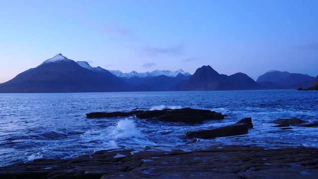 Evening in Elgol bay on west coast of the Isle of Skye in Scotland during an windy sunset . Snowy mountains on Soay island above sea of the Hebrides, waves crashing against to boulders and sharp rock 
