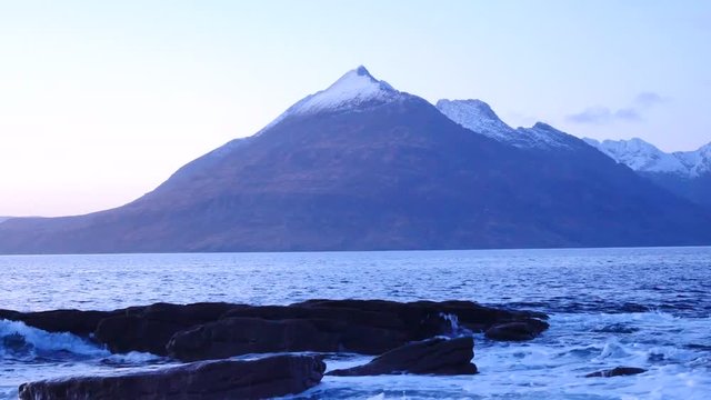 Evening in Elgol bay on west coast of the Isle of Skye in Scotland during an windy sunset . Snowy mountains on Soay island above sea of the Hebrides, waves crashing against to boulders and sharp rock 
