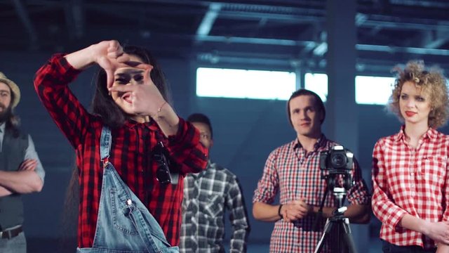 Young beautiful asian woman stepping out and making frame gesture with fingers while group of young filmmakers shooting video with dslr camera