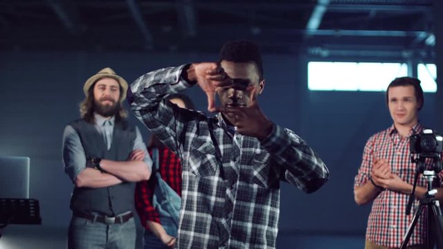 Young african american man stepping out and making frame gesture with fingers while group of young filmmakers shooting video with dslr camera