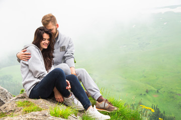Naklejka na ściany i meble Happy young couple in love sitting on a mountain, hugging and looking away, love and feelings concept