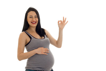 happy young brunette pregnant girl with hand on her belly showing OK isolated on white background