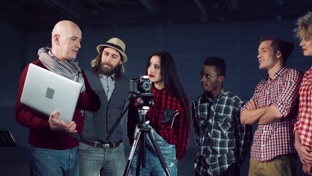 Bold man producer with laptop in hand talking to crew of young people filmmakers standing near DSLR camera on tripod in dark empty hall. Close shot
