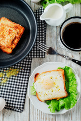 a delicious and hearty breakfast, fried eggs in a white toast with spices, peppers, lettuce, a cup of black coffee and milk on a wooden background 