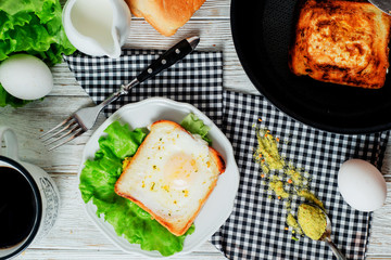 a delicious and hearty breakfast, fried eggs in a white toast with spices, peppers, lettuce, a cup of black coffee and milk on a wooden background 