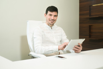 A young business man facing camera, smiling while working tablet - I pad. A clean office desktop sitting in a white chair