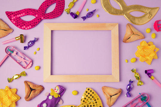 Purim holiday concept with wooden frame, carnival mask and hamans ears cookies on purple background. Top view from above. Flat lay