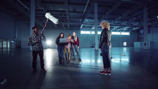 Group of young people with camera on tripod, laptop and boom mic setting gear for shooting scene with man in hat and caucasian young girl in leather jacket standing and playing role or giving