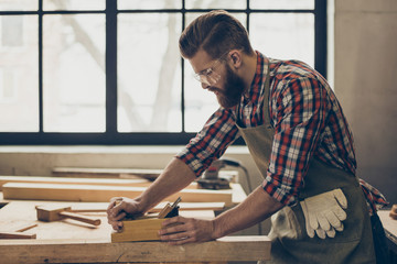 side view photo of stylish handsome cabinetmaker work with jointer and plank. Man with brutal hairstyle, beard and glasses