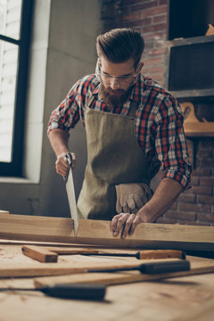 young stylish cabinet-maker with  glasses and hairstyle. Strong  handsome craftsman holding saw and wood blank at workplace