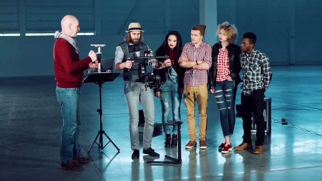 Group of filmmakers using handheld camera gyro stabilizing gimbal. Crew of young men and women watching the director showing cameraman some moves standing in empty studio hall