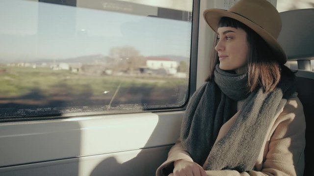 Beautiful single girl in beige hat travels in train, looks out the window on country landscape and smiles thinking about something, soft morning sunlight shines through window