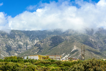 Mountainous nature of Crete Island in Greece. Low clouds hide tops of mountains under blue sky. Houses and typical wooden electric poles with wires under voltage.