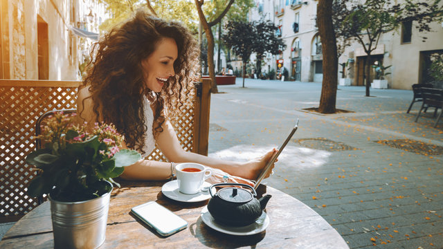 Young curly woman talking in video chat with her friend via digital tablet while sitting in street cafe and drink delicious black tea, laughing girl watching comedy movie on touch pad during breakfast