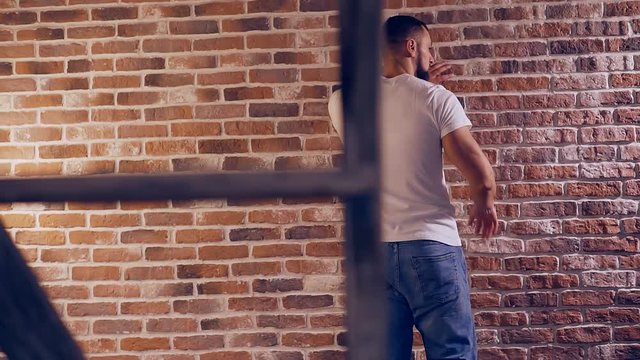 A man in jeans and a white t-shirt from the back takes place in a country house to the brick wall and thinks.