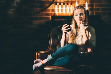 Girl in a cozy dark room holding a phone and searches the interenet. Blonde and warm lamp light