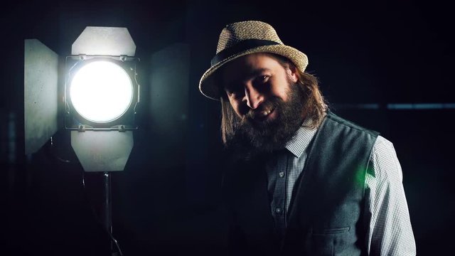 Bearded man in hat and vest standing at studio light stand with switcher in hand, turning it on and checking and smiling, looking at camera