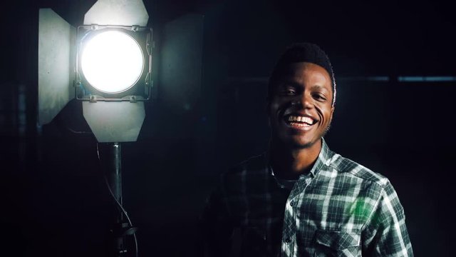 Young African-American man in darkness coming close to camera, then studio light stand is being turned on making man happy and smiling. Looking at camera