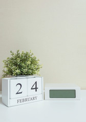 White wooden calendar with black 24 february word with clock and plant on white wood desk and cream wallpaper textured background in selective focus at the calendar