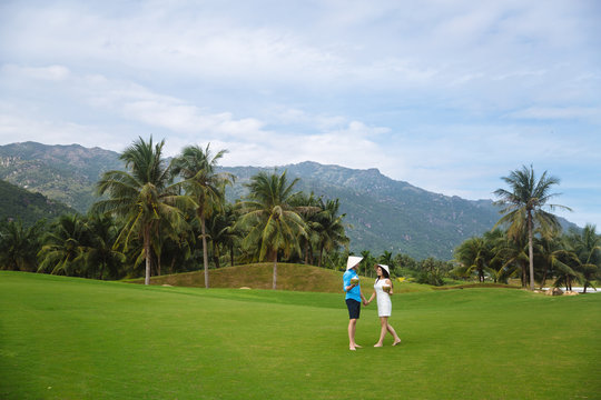 Young beautiful couple in love holding coconut in hands and walking at golf course in summer. man wear the blue shirt and the girl in a white dress and vietnamese hats. Concept of honeymoon