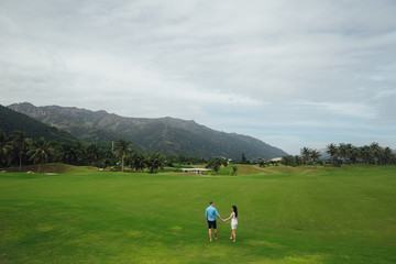 Young beautiful couple in love holding hands and walking at golf course in summer. man wear the blue shirt and the girl in a white dress. Concept of honeymoon