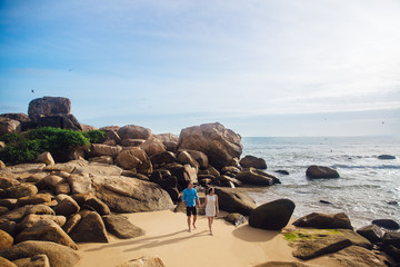 Young couple in love holding hands, walking on a sea beach in summer. The man in the blue shirt and the girl in a white dress with a wreath in her hair