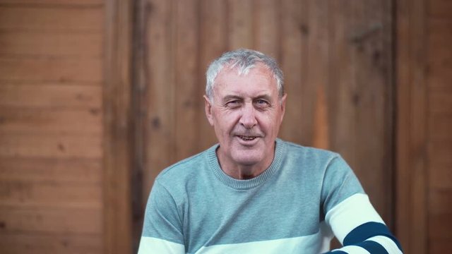 Portrait of old man sitting on the porch of the house. Pensioner looking to the camera, smiling and smoking. 4K
