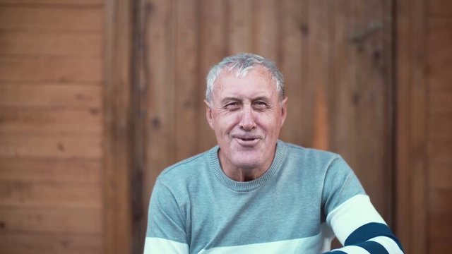 Old man sitting on the porch of the house. Grandfather making funny face and granddaughter running to him, hugs. 4K
