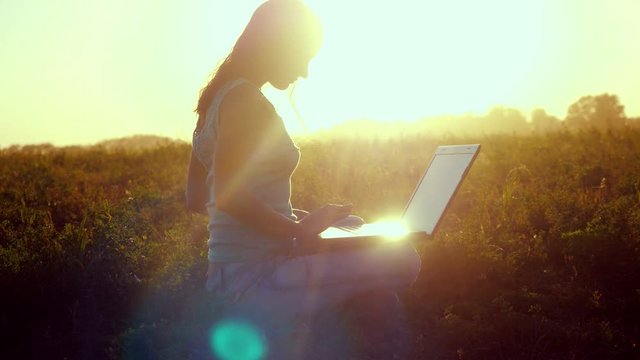 Young brunette woman typing on a laptop outdoors at beautiful sunset sitting on the grass with amazing lense flare effects in summer time. 3840x2160