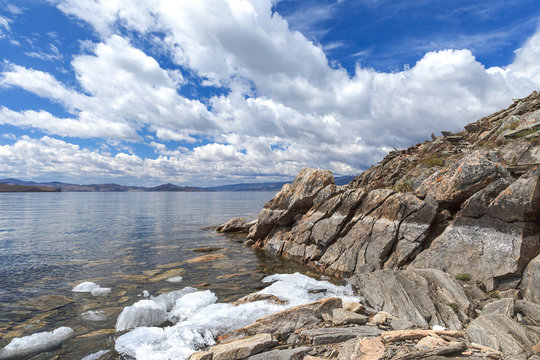 Landscape with textured rock, white clouds and white melting ice along coastline in spring day