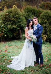 Groom in plaid blue jacket holds bride's waist standing with her on green lawn