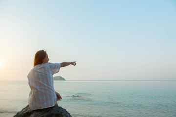 young woman sitting on a rock enjoying the beach and pointing to sky