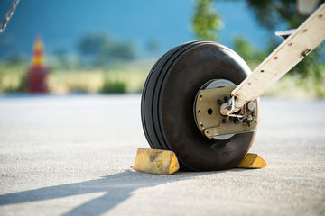 A rear landing gear and wheel chocks of a small aircraft on the ground with blurry nature and traffic cone in the background.