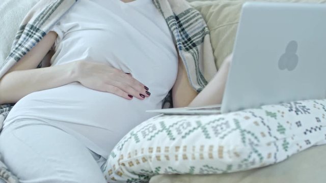 Tilt up of smiling pregnant woman stroking her belly while sitting on sofa at home and surfing the Net on laptop